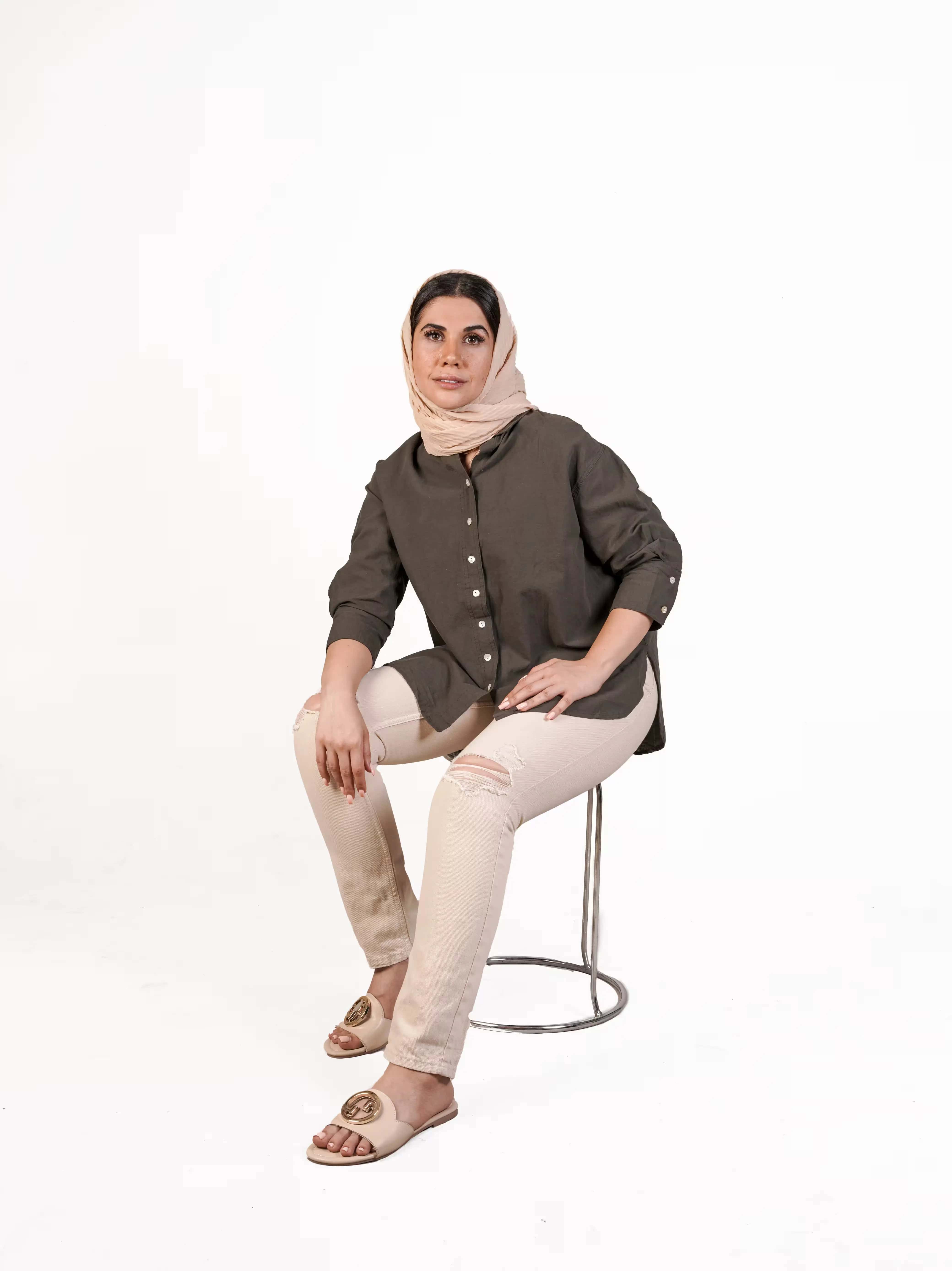 شومیز لینن زنانه وارداتی با رنگ خاکی و آستین سه ربع، یقه کلاسیک و دکمه‌دار، تنخور آزاد، ست شده با شلوار اسکینی کرم و شال روشن روی مدل نشسته روی صندلی، مناسب استایل روزمره و رسمی.